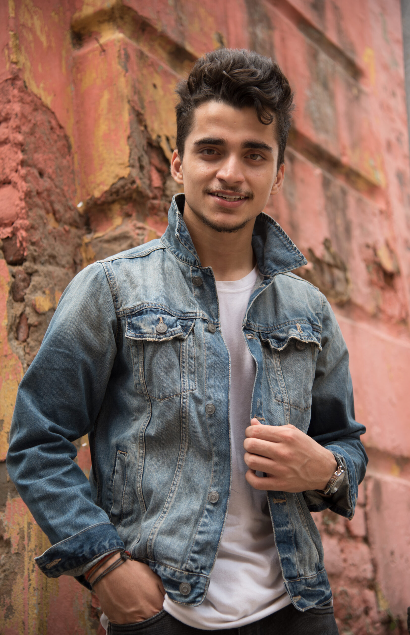 An attractive South Asian male posing in front of a brick wall wearing a jean jacket